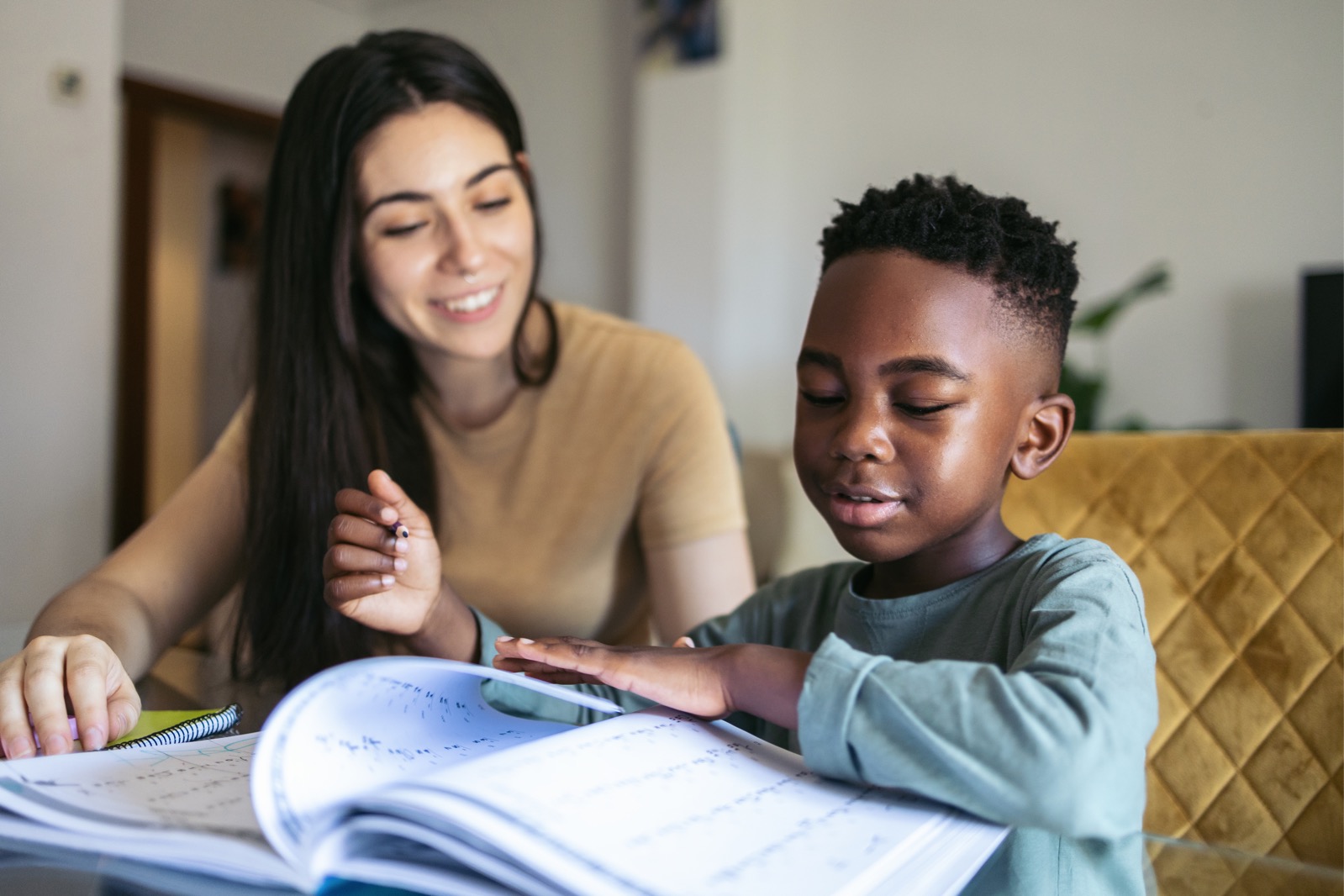 Little boy doing homework at home with female adult. They are sitting at the table in the living room with their notebooks and school books.
